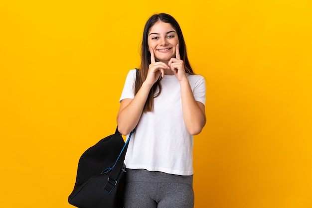 Young sport woman with sport bag isolated on yellow wall smiling with a happy and pleasant expression