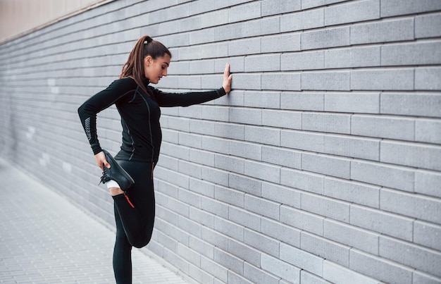 Young sportive girl in black sportswear outdoors doing stretching near gray wall