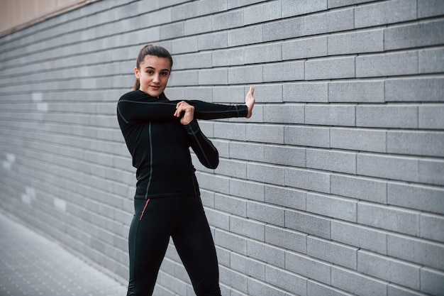 Young sportive girl in black sportswear outdoors doing stretching near gray wall