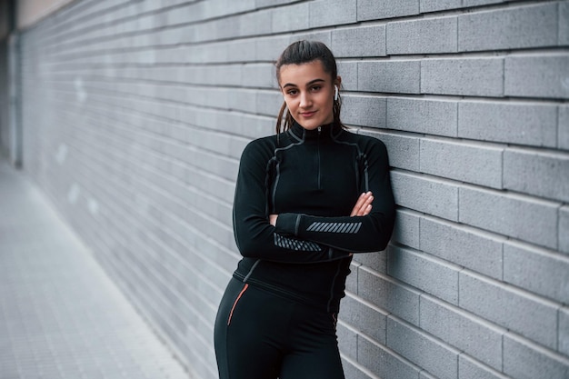 Young sportive girl in black sportswear outdoors near gray wall