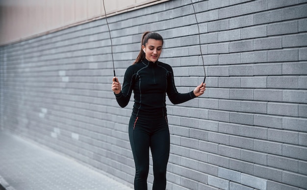 Young sportive girl in black sportswear practicing with jump rope outdoors near gray wall