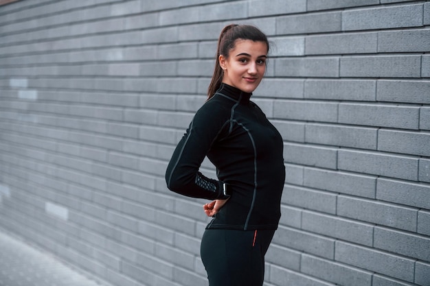 Young sportive girl in black sportswear standing outdoors near gray wall