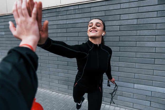 Young sportive girl in black sportswear standing with jump rope in hands outdoors near gray wall and giving high five