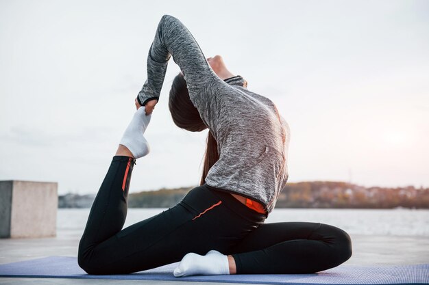 Young sportive girl doing yoga exercises on fitness mat outdoors near lake at daytime