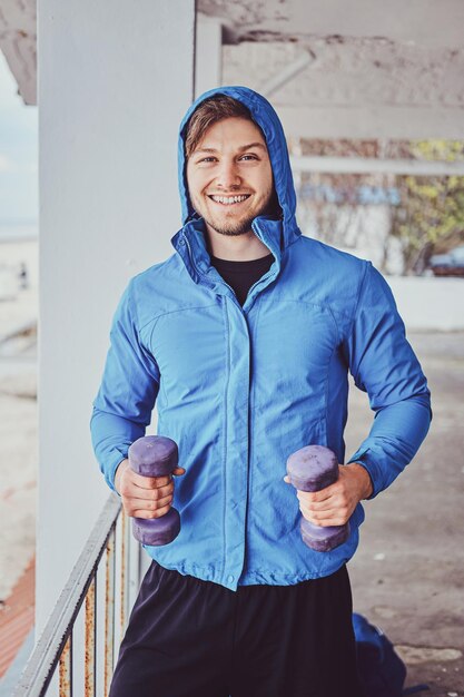 Young sportive man is doing exercises outside with dumbbells while posing for photographer.