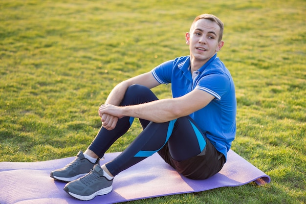 Young sportive man sitting on training mat in morning field outdoors.