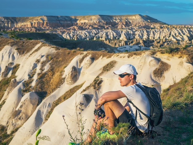 Young sportive man in white cap with backpack sitting on the peak of sandstone rock