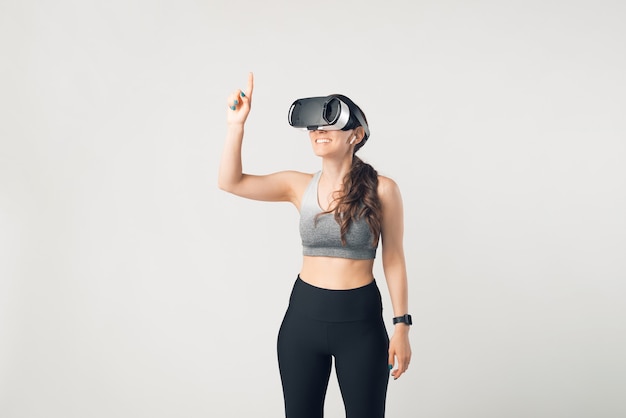 Young sportive woman is wearing some VR glasses and sportswear over white background.