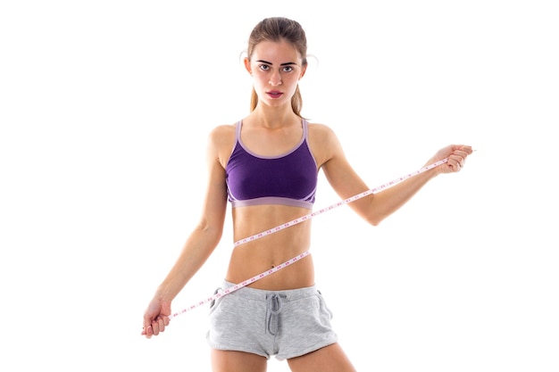 Young sportive woman in purple sports top with ponytail using centimeter on white background in studio