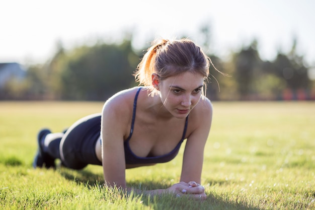 Young sportive woman in sports clothes training in field at sunrise. Girl standing in plank position on grass in a park.