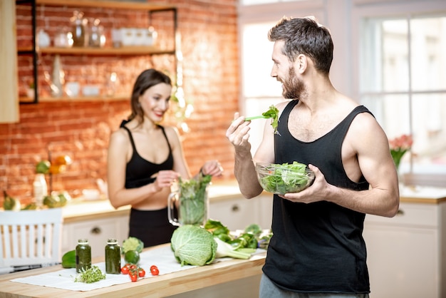 Young sports couple having snack with healthy salad and green smoothie on the kitchen at home