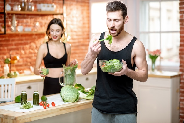 Giovani coppie sportive che fanno uno spuntino con insalata sana e frullato verde in cucina a casa
