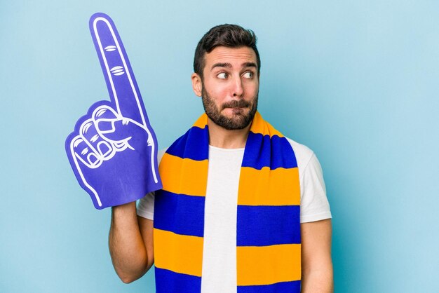 Young sports fan man isolated on blue background confused feels doubtful and unsure