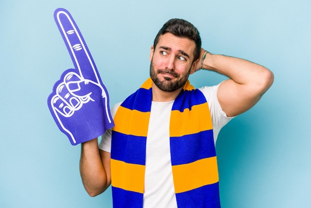 Young sports fan man isolated on blue background touching back of head thinking and making a choice