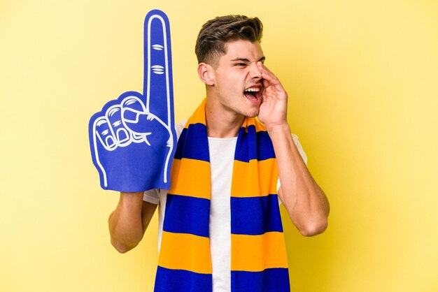 Young sports fan man isolated on yellow background shouting and holding palm near opened mouth.