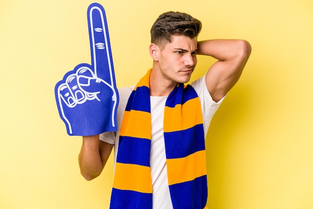Young sports fan man isolated on yellow background touching back of head, thinking and making a choice.