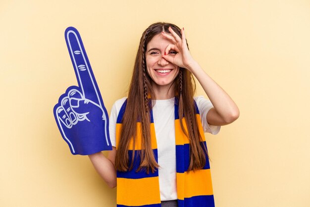 Young sports fan woman isolated on yellow background excited keeping ok gesture on eye