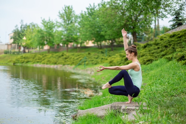 A young sports girl practices yoga on a green lawn by the river