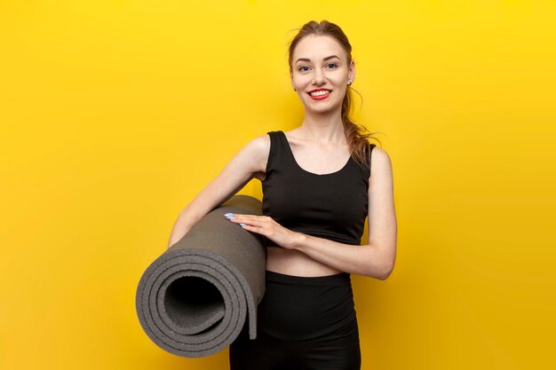 Молодая спортивная девушка в спортивной одежде с ковриком для йоги улыбается на желтом изолированном фоне
