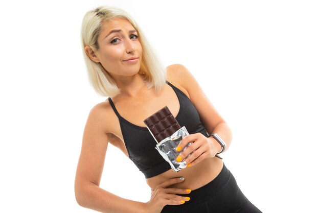 A young sports girl with blonde hair in a black sports top and black leggings holds chocolate and think