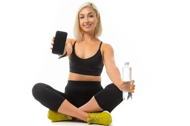 Una giovane ragazza sportiva con i capelli biondi e la manicure brillante in un top sportivo nero e leggings tiene una bottiglia d'acqua e mostra un telefono nero.