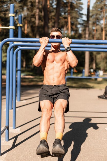 Young sports man athlete with a sexy muscular torso hangs on horizontal bars and trains in the park on a sunny day Healthy and workout lifestyle