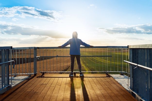 Young sports man watch the agricultural fields over bridge handrail. Dramatic evening scene, rays of the setting sun.