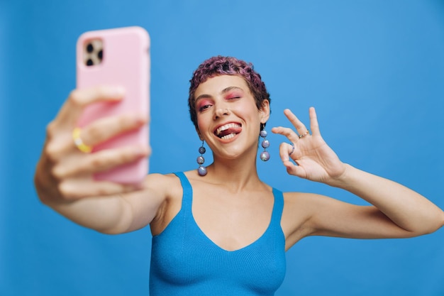 Young sports woman fashion blogger takes a picture of herself on the phone in blue sportswear smiling and showing her tongue on a blue monochrome background