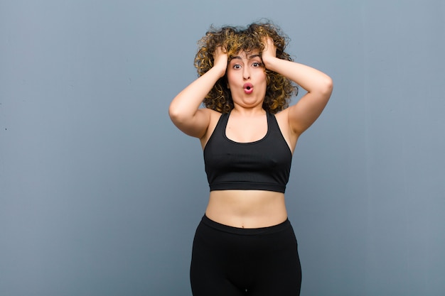 Young sports woman looking excited and surprised, open-mouthed with both hands on head, feeling like a lucky winner over gray wall