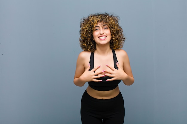 Young sports woman looking happy, surprised, proud and excited, pointing to self on gray wall