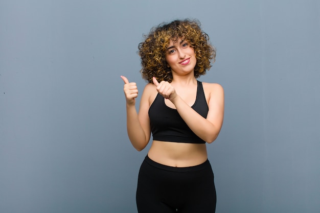 Young sports woman smiling cheerfully and casually pointing to copy space on the side, feeling happy and satisfied against gray wall