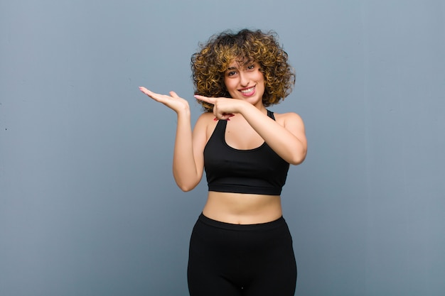 Young sports woman smiling cheerfully and pointing to copy space on palm on the side, showing or advertising an object against gray wall