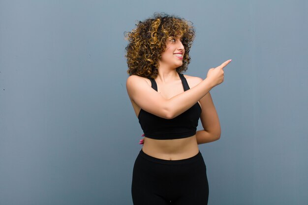 Young sports woman standing and pointing to object on copy space, rear view against gray wall