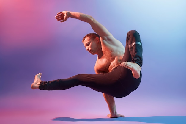 Young sportsman acrobat or gymnast posing against neon wall, showing his power, stands on one hand, wearing black trousers, demonstrates his sports achievements.