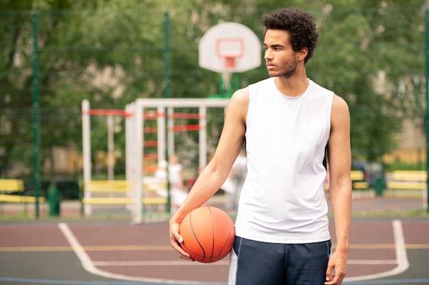 Foto giovane sportivo in activewear che osserva da parte pur avendo una pausa tra gli allenamenti di basket sul campo