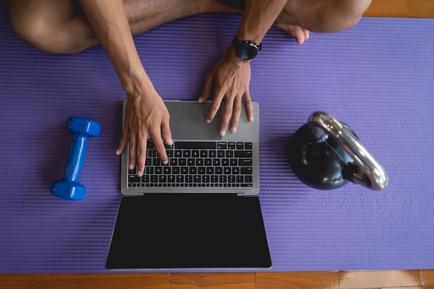 Young sportsman sitting on purple yoga mat and doing exercise\
sports workout online and looking video streaming on laptop indoor\
at the fitness gym online fitness training from gym concept