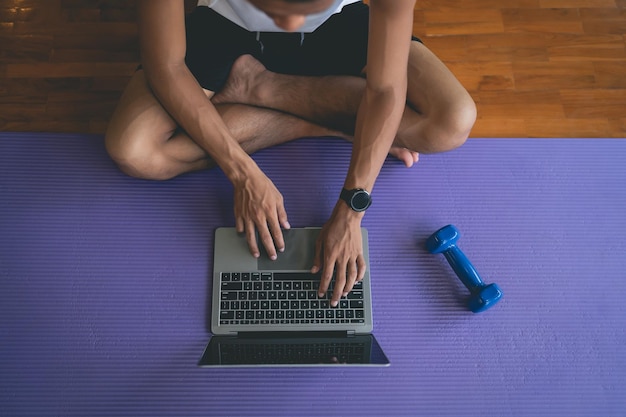 Young sportsman sitting on purple yoga mat and doing exercise sports workout online and looking video streaming on laptop indoor at the fitness gym Online fitness training from gym concept