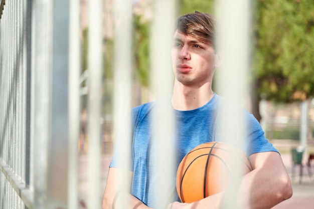 Young sportsman with a basketball in his hand leaning against a
metal fence on a basketball court