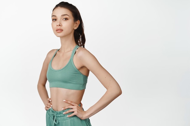 Young sportswoman in activewear looking confident holding hands on waist and staring at camera motivated for good workout standing over white background