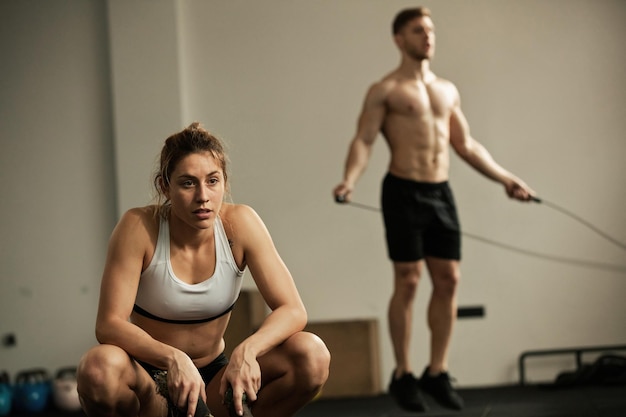 Young sportswoman crouching while feeling exhausted after exercising with battle ropes in gym Man skipping rope in the background