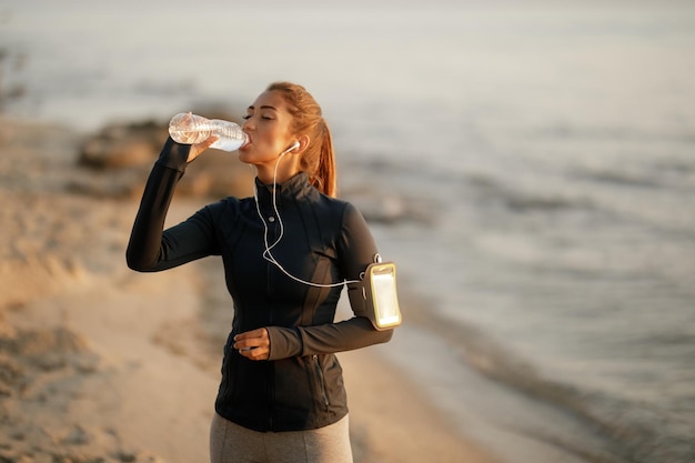 Молодая спортсменка пьет воду из бутылки, отдыхая от утренней пробежки на пляже