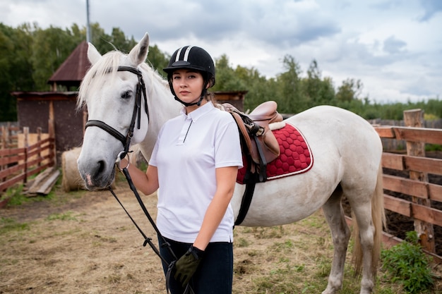 カメラの前で白い純血種の競走馬によって立っている乗馬服の若いスポーツ選手