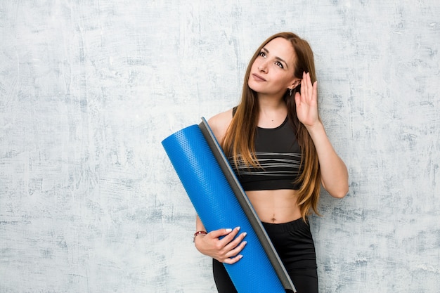 Young sportswoman holding a mat trying to listening a gossip