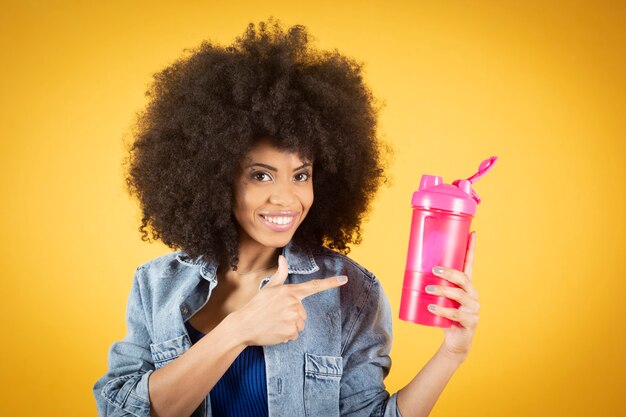 Young sportswoman relaxing on the floor and drinking smoothie on yellow background, happy mixed african