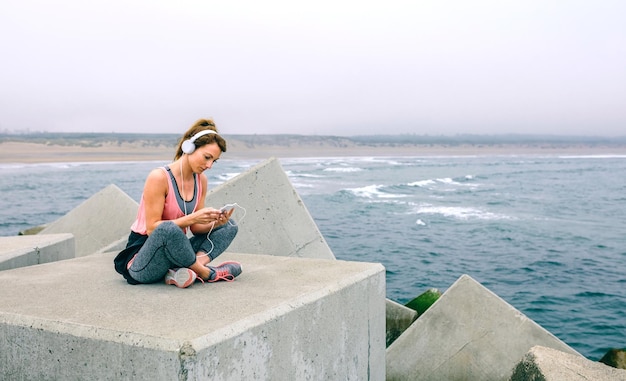 Young sportswoman with headphones sitting by sea pier