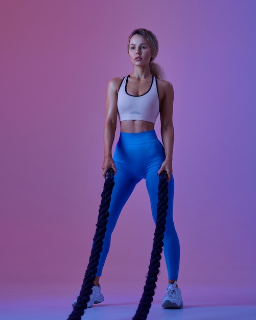 Young sportswoman with ropes poses in studio, neon background