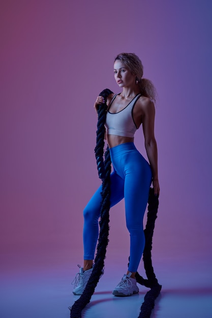 Young sportswoman with ropes poses in studio, neon background