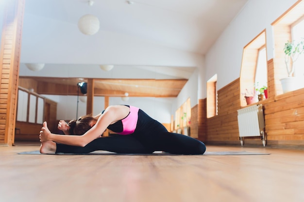 Young sporty attractive woman practicing yoga doing Revolved Head to Knee Forward exercise Parivrtta Janu Sirsasana pose working out wearing sportswear black pants indoor close up yoga studio