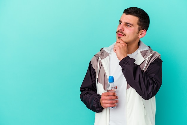 Young sporty caucasian man drinking water isolated on blue looking sideways with doubtful and skeptical expression.