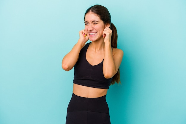 Young sporty caucasian woman isolated on blue background covering ears with hands.